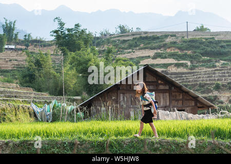 Sapa, Vietnam - Mai 2019: Mädchen aus der ethnischen Gruppe der Hmong in traditioneller Kleidung trägt Baby im Tragetuch in Lao Cai Provinz. Stockfoto