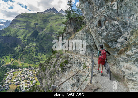 Aktive Senior-Frau, die ihr Elektro-Mountainbike auf einem schmalen Mountainbike-Trail hoch über Zermatt, dem berühmten Touristenziel im südwesten, schiebt Stockfoto