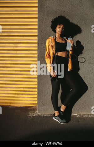Gesunde junge Frau mit Wasserflasche stehen gegen eine Wand. Die volle Länge der sportliche junge Frau entspannend nach dem Ausführen der Übung im Freien.