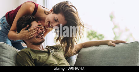 Lächelnden jungen Frau die Augen ihres Partners sitzen auf einem Sofa. Junge Frau überraschend Mann im Wohnzimmer zu Hause. Stockfoto