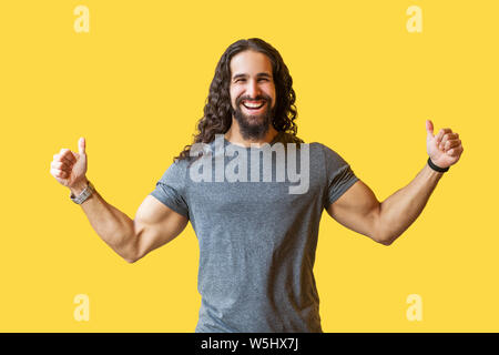 Portrait von Happy bärtiger junger Mann mit langen, lockigen Haar grau T-Shirt mit Daumen hoch stehen und an der Kamera mit toothy Lächeln und zuversichtlich Stockfoto