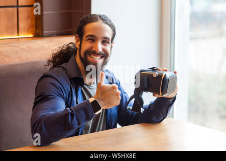 Portrait von Happy bärtiger junger erwachsener Mann in lässigen Stil im Cafe sitzen, Holding vr und Daumen oben am Simulator mit glücklichen Gesicht, c Stockfoto