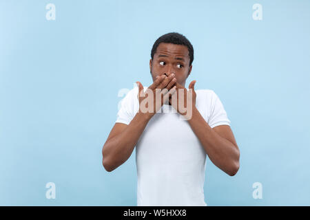 Halbe Länge close up Portrait von jungen afrikanischen - amerikanische Mann im weißen Hemd auf blauem Hintergrund. Menschliche Gefühle, Mimik, Ad-Konzept. Schockiert über den Mund, die durch seine Hände. Stockfoto