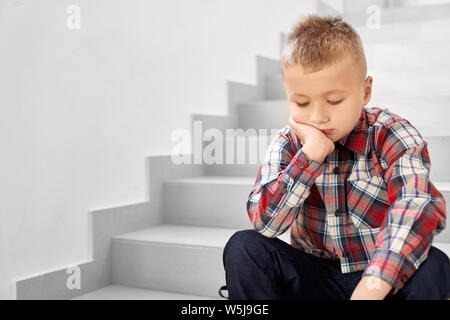 Eine leider Schule Junge in kariertes Hemd sitzt auf der Treppe im Flur der Schule und weinte. Kleine Schule junge schiefen Kopf durch Hände und Denken. Konzept der bed Noten in der Schule. Stockfoto