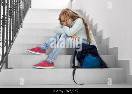 Seitenansicht des blonden Mädchen sitzen auf der Treppe ein- und ausblenden von gekreuzten Händen, an der Kamera schaut. Leider Mädchen weinend auf Korridor, denn Bett Markierung an der Lektion. Einsame Schüler mit Bett Stimmung. Stockfoto