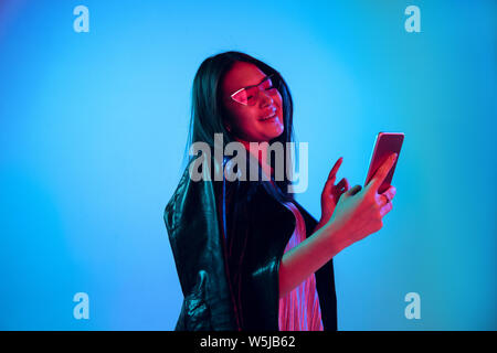 Schöne weibliche Brustbild auf Blau studio Hintergrund im Neonlicht isoliert. Junge emotionale Frau. Menschliche Gefühle, Mimik Konzept. Mit Smartphone und kabellosen Kopfhörern. Stockfoto