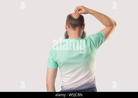 Rückseite Porträt der glatzköpfige Mann mittleren Alters mit Bart in hellem Grün t-shirt stehend, den Kopf kratzen oder Denken über etwas. indoor Studio s Stockfoto