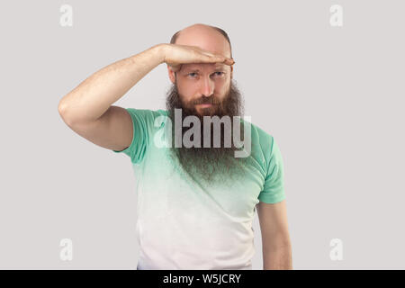 Portrait von aufmerksamen Kahl im mittleren Alter Mann mit langem Bart in hellem Grün t-shirt stehen mit Hand auf Stirn und weit schauen, um etwas zu finden. Stockfoto