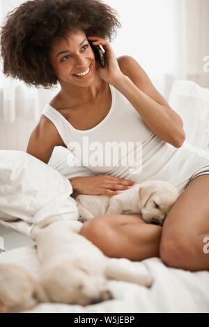 Junge Frau im Gespräch über Ihr Handy beim Sitzen auf dem Bett mit Schlaf Labrador Welpen. Stockfoto