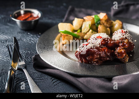 Gegrilltes Hähnchen shins in Tomatensauce mit Chili und gebackene Kartoffeln auf einer Metallplatte auf einem dunklen Hintergrund. Das Essen im Restaurant. Nahaufnahme Stockfoto