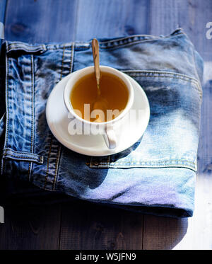 Becher gefüllt mit kochendem Wasser und Tee Beutel auf Blue Jeans Hintergrund. Prozess Kaffee brühen in Keramik Becher. Pflanzliche grün oder schwarz vollständige Blatt. Gesunde Gewohnheiten. Tea time Konzept. Tasse Tasse heißes Wasser und Beutel mit Tee. Stockfoto