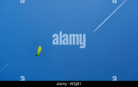 Ein Gleitschirm mit einem gelb-grün parashute und ein Flugzeug verlassen eine weiße Spur auf den blauen Himmel. Stockfoto