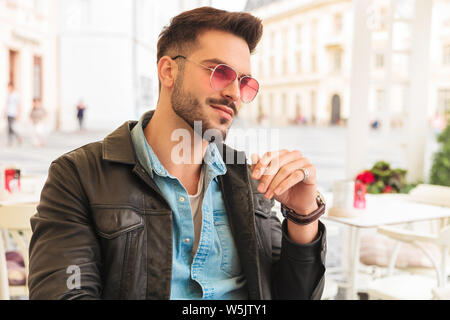 Portrait von neugierig und lächelnd Mode Mann tragende rote Sonnenbrille und Lederjacke sitzen im Stadtzentrum, an einem Tisch und Suchen auf Seite Stockfoto