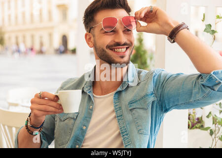 Neugierig und lächelnd legerer Mann zur Festsetzung Sonnenbrille Getränke Kaffee, während Sie an einem Tisch in der Stadt saßen und zu Seite, portrait Bild Stockfoto
