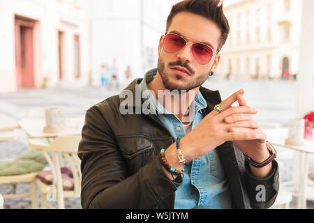 Mode Mann Sonnenbrille tragen und halten Handflächen zusammen sieht, beim Sitzen am Tisch im Stadtzentrum zu Seite Stockfoto