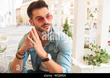 Neugierig legerer Mann tragende rote Sonnenbrille holding Handflächen zusammen, während am Tisch in der Stadt sitzen schaut zur Seite, portrait Bild Stockfoto