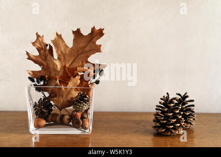 Stillleben mit herbstlichen Home Decor von golden und Braune trockene Blätter, Kegel und Eicheln in modernen Glas Vase und zwei Kegel auf Holztisch, kopieren Raum Stockfoto