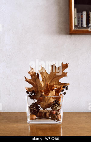 Herbstliche Home Decor mit goldenen und braunen Blätter, Zapfen und Eicheln in modernen Glas Vase auf dem Tisch, Kopieren, teilweise sichtbar Spiegel mit Bücherregal Stockfoto