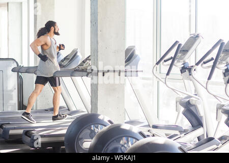 Seitenansicht junger Erwachsener sport Mann mit Langen lockigen Haare Training im Fitnessstudio allein. Stattliche männliche Jogging auf Laufband und Fenster. Hallenbad, Kur Stockfoto