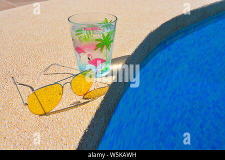 Pool Tag, Sommer Fun Vibes. Paar Metall eingefasste gelbe Sonnenbrille und einem funkelnden kaltes Getränk in einem Plastikbecher am Pool. Stockfoto