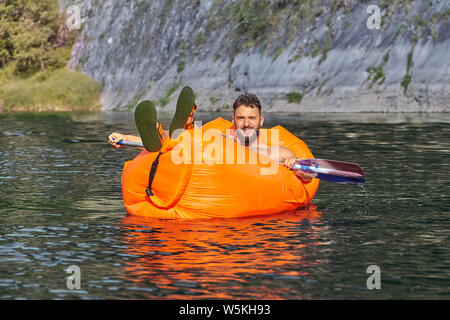 Junge Bartgeier kaukasischen Mann liegen auf dem Orange aufblasbarer Liegestuhl mit Schiff in seiner Hand, während die Sofa schwimmen auf den ruhigen Fluss ist mit Einfassung Stockfoto