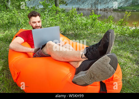 Junge Bartgeier Geschäftsmann mit Notebook ist auf der Orange aufblasbarer liege bei eco - Tourismus in der Nähe des Flusses. Stockfoto