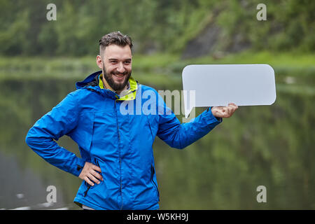 Junge attraktive Bärtigen kaukasischen Mann mit leeren Blatt steht in der wilden Natur in der Nähe des Flusses in der blauen Jacke allein und lächelnd. Stockfoto