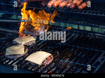 Zangen sind herauf einen Hamburger wie Flammen aus der Kohle umgeben, und Cheeseburger und sind alle kochen Hot Dogs auf dem Grill. Stockfoto