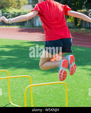 Ein High School Jugendmädchen über zwei Meter hohen Gelbe Mini Hürden während der Leichtathletik Praxis springen ist auf einem grünen Rasenfeld. Stockfoto