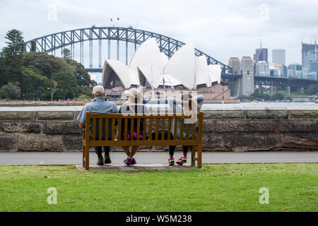 Drei Leute sitzen auf einer Bank mit Blick auf das Sydney Opera House und der Sydney Harbour Bridge, von der Farm Cove, Sydney Stockfoto