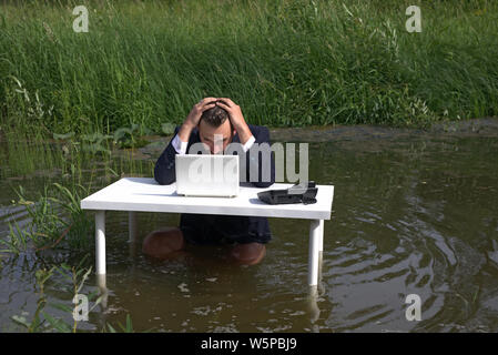 Junger bärtiger Mann, Mann seinen Kopf Holding frustriert. Versucht, sich auf die Arbeit zu konzentrieren. von der Arbeit müde. Am Tisch, im Sumpf, im Wasser Stockfoto
