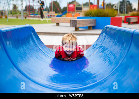 Ein 2-jähriger Junge geht nach unten eine blaue Folie auf seinem Bauch, Spaß an einen neuen Spielplatz, in Canterbury, Neuseeland Stockfoto