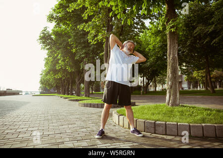 Ältere Menschen als Runner an der Hauptstraße der Stadt. Kaukasische männliches Modell joggen und cardio Training in Sommermorgen. Dabei Stretching Übungen in der Nähe der Wiese. Gesunder Lebensstil, Sport, Aktivität Konzept. Stockfoto