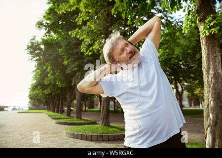 Ältere Menschen als Runner an der Hauptstraße der Stadt. Kaukasische männliches Modell joggen und cardio Training in Sommermorgen. Dabei Stretching Übungen in der Nähe der Wiese. Gesunder Lebensstil, Sport, Aktivität Konzept. Stockfoto