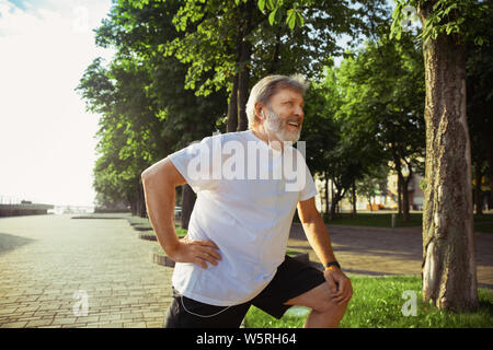 Ältere Menschen als Runner an der Hauptstraße der Stadt. Kaukasische männliches Modell joggen und cardio Training in Sommermorgen. Dabei Stretching Übungen in der Nähe der Wiese. Gesunder Lebensstil, Sport, Aktivität Konzept. Stockfoto