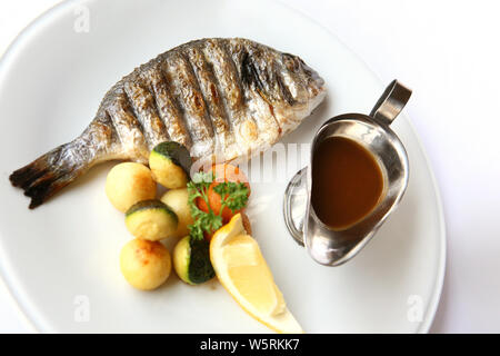 Gegrillter Fisch auf einem weißen Teller mit Gemüse, Zitrone und Soße auf weißem Hintergrund Stockfoto