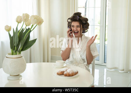Schöne junge lächelnde brünette Frau in Lockenstäbe sprechen auf dem Smartphone beim trinken Kaffee und essen Croissants zum Frühstück im hellen Stockfoto