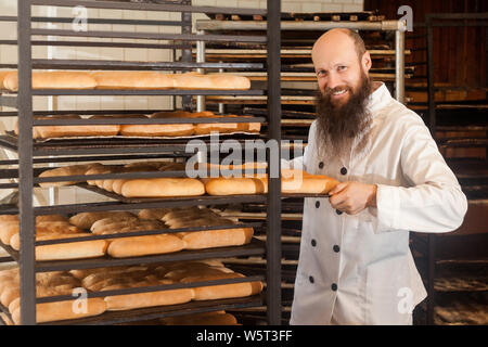 Portrait von Freude junger Erwachsener Bäcker mit langen Bart in weiße Uniform stehend in der Nähe der Regale voll mit frisch gebackenen Brot in seinen Arbeitsplatz, ICH Stockfoto