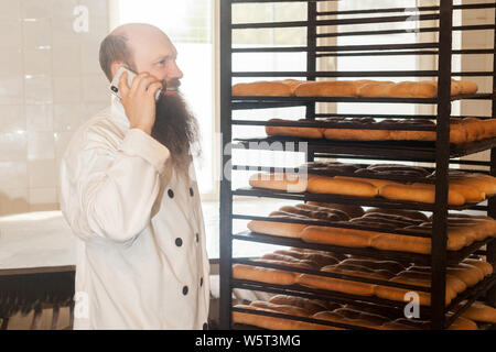 Portrait von glücklichen jungen Erwachsenen Geschäftsmann Bäcker mit langen Bart in weiße Uniform stehend in Bäckerei und haben online Bestellung per Telefon, sprechen an cellpho Stockfoto