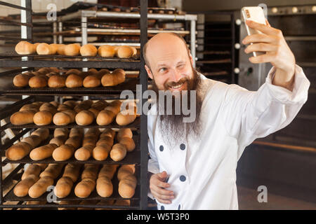 Portrait von fröhlicher junger Erwachsener blogger Bäcker mit langen Bart in weiße Uniform stehend in Fabrik und selfie auf Regalen mit frischem Brot zurück Stockfoto