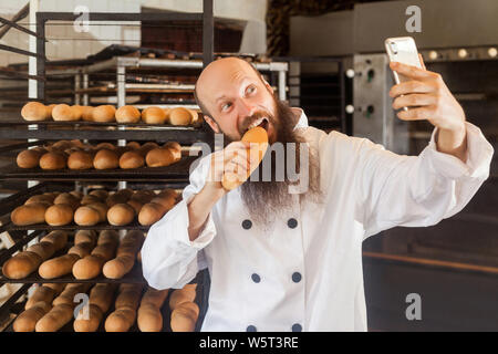 Portrait von hungrigen jungen Erwachsenen blogger Bäcker mit langen Bart in weiße Uniform stehend in Fabrik und selfie auf Regalen mit frischem Brot backgr Stockfoto