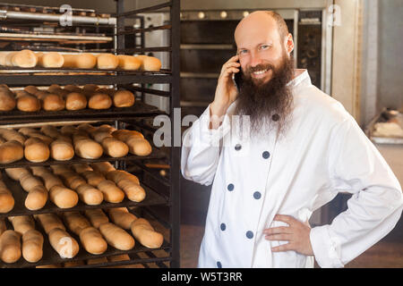 Portrait von glücklichen jungen Erwachsenen Geschäftsmann Bäcker mit langen Bart in weiße Uniform stehend in Bäckerei und haben online Bestellung per Telefon, sprechen an cellpho Stockfoto