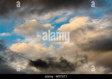 Golden Fluffy Clouds von verschwindenden Strahlen bei Sonnenuntergang und dunklen Gewitterwolken schwimmend über sonnigen blauen Himmel Sommer Wetter zu ändern beleuchtet. Stockfoto
