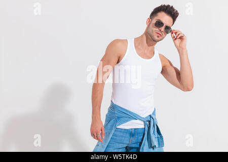 Portrait von junger Mann in Unterhemd zur Festsetzung seiner Sonnenbrille beim Stehen auf weißen Hintergrund und lehnte sich zur Seite Stockfoto