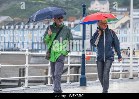 Aberystwyth, Wales, UK. 30. Juli 2019. Urlauber in ihre regenjacken und Schirme zu Fuß entlang der Promenade in Aberystwyth im Regen wie ein Gürtel von thundery und sehr nassem Wetter breitet sich über die zentralen Teile des UK, damit besteht die Gefahr von Überschwemmungen und gefährlichen Fahrbedingungen.. Foto: Keith Morris/Alamy leben Nachrichten Stockfoto