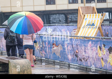 Aberystwyth, Wales, UK. 30. Juli 2019. Urlauber in ihre regenjacken und Schirme zu Fuß entlang der Promenade in Aberystwyth im Regen wie ein Gürtel von thundery und sehr nassem Wetter breitet sich über die zentralen Teile des UK, damit besteht die Gefahr von Überschwemmungen und gefährlichen Fahrbedingungen.. Foto: Keith Morris/Alamy leben Nachrichten Stockfoto