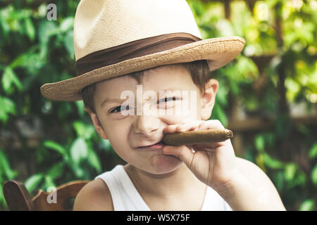 Schöne junge spielt Nachahmung ein Erwachsener, raucht eine Zigarre. Funny kid macht Flächen durch rümpfte die Nase, während Sie eine Zigarre in der Hand. Schließen Stockfoto