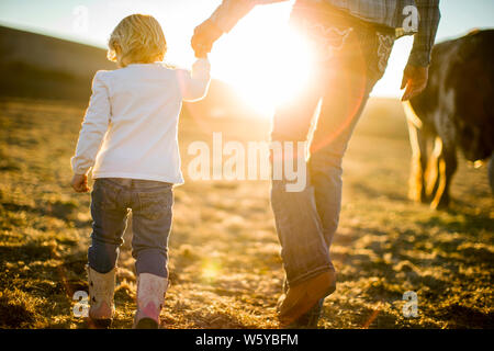 Bäuerin und ihren Toddler Tochter gehen Hand-in-Hand durch die Ranch Paddocks bei Sonnenuntergang. Stockfoto