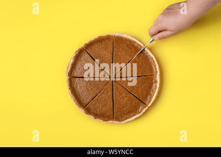 Schneiden eine Torte Konzept. Frau Hand in Scheiben schneiden eine Kürbistorte auf gelbem Hintergrund. Blick von oben auf die köstliche traditionelle Thanksgiving Nachtisch. Stockfoto