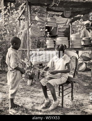 1940ER AFRICAN AMERICAN JUNGE STEHEND BEOBACHTEN MÄDCHEN WEBKORB AN STAND MIT MEHREREN KÖRBEN ZUM VERKAUF SOUTH CAROLINA USA - N1202 HAR001 HARS 1 JUGENDSTIL LIFESTYLE GESCHICHTE FRAUEN BRÜDER LANDHAUSLEBEN VEREINIGTE STAATEN KOPIEREN RAUM FREUNDSCHAFT HALBEN LÄNGE HANDWERK TRADITIONELLE VEREINIGTE STAATEN VON AMERIKA MÄNNER KÜNSTLER HANDWERK GESCHWISTER SCHWESTERN B&W KÜNSTLER VERKAUFEN AFROAMERIKANISCHE AFROAMERIKANISCHE SCHWARZE ETHNIE CAROLINA LOKAL BERUFE GESCHWISTER SÜD KUNST VERKAUFEN TRADITIONEN KÖRBE KREATIVITÄT HANDWERK JUVENILES GULLAH PRE-TEEN MÄDCHEN STRASSENRAND SC MEHRERE SOUTH CAROLINA TALENT ZWEISAMKEIT Stockfoto